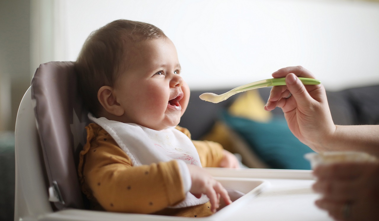 A baby in a highchair opens it's mouth to be fed from a baby spoon.