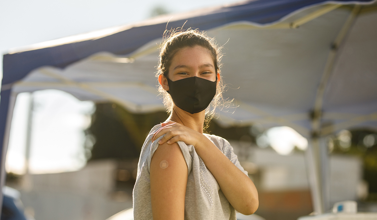 A teenage girl wearing a face mask shows the band-aid from her vaccine shot. 