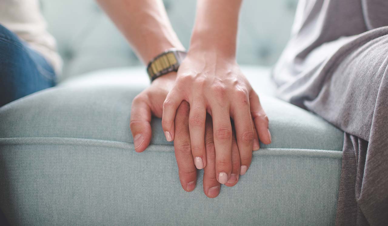 Sitting on a couch, a couple holds each others hands.