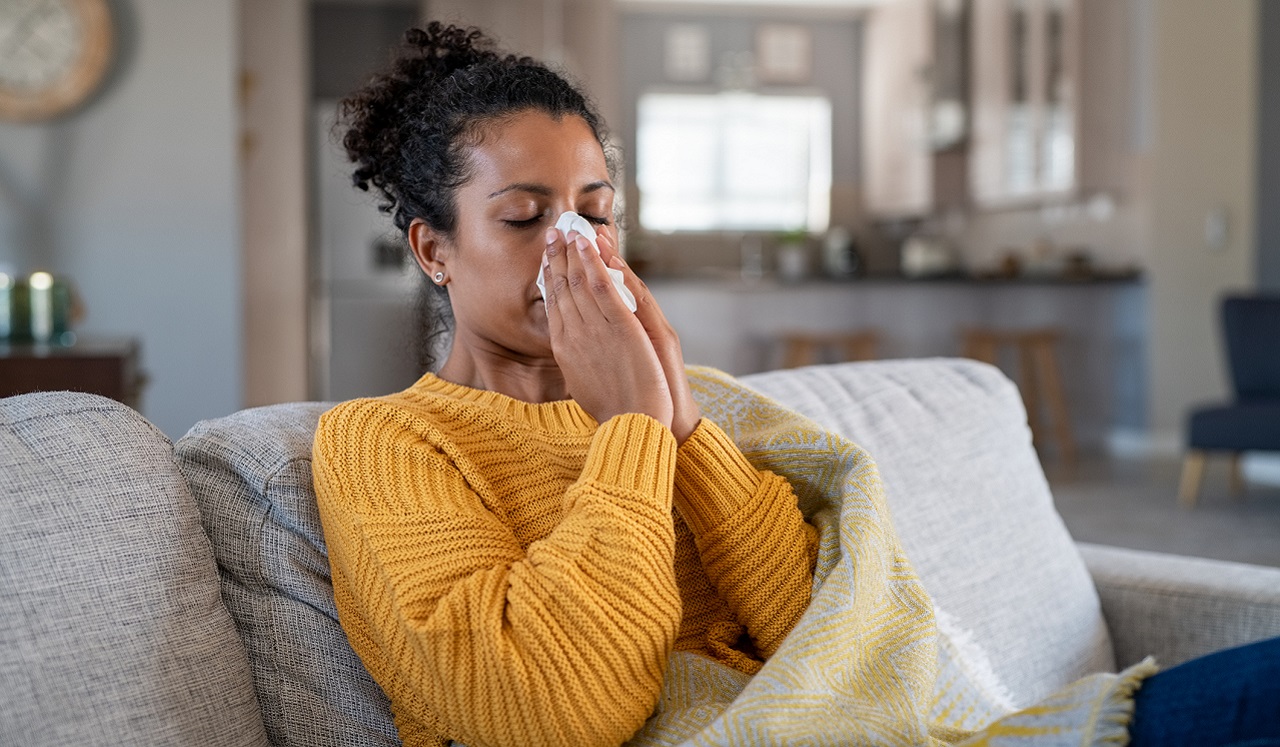 A woman holds a tissue to her nose as she sits on the couch.