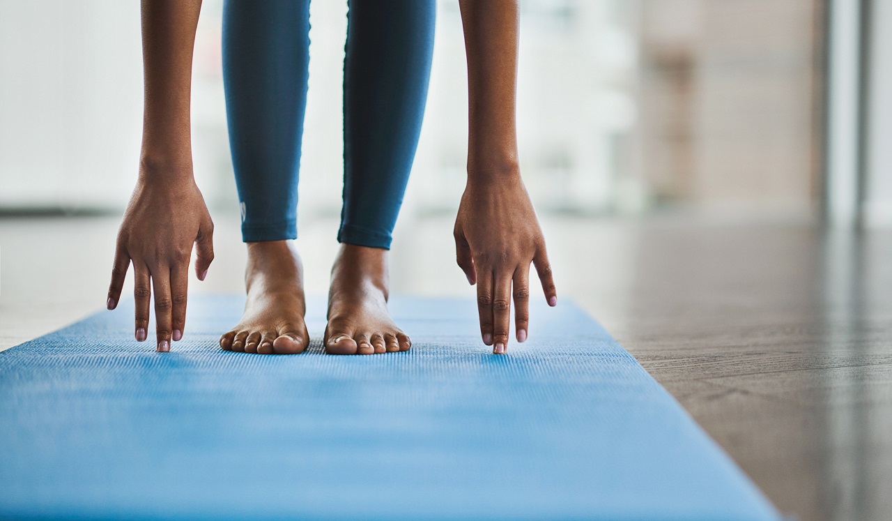 From the top of a blue yoga mat, we see a woman standing as she reaches her finger tips to the matt.