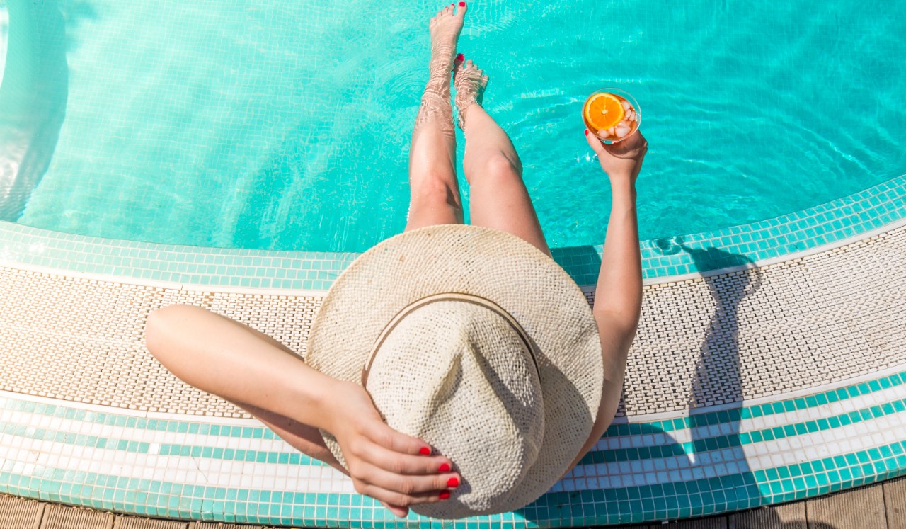 A woman in a brimmed hat sits pool side with her feet in the water.
