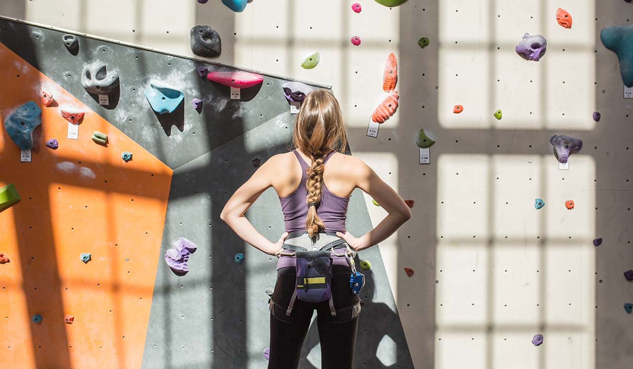 A woman in climbing gear looks up at a climbing rock wall.
