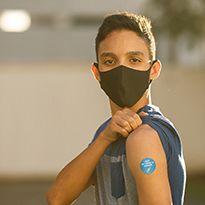 A young boy donning a mask rolls up his sleeve to show his vaccine injection site.