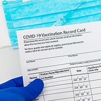 A gloved hand holds a vaccination record card; in the background is a syringe and a medical face mask.
