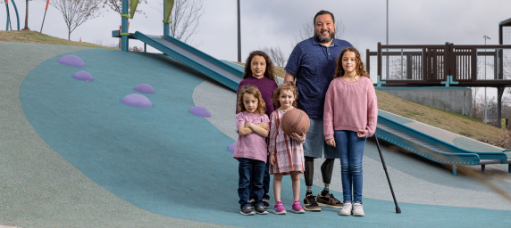 TIRR Memorial Hermann Osseointegration patient, Alex, stands with his four daughters at a play ground.
