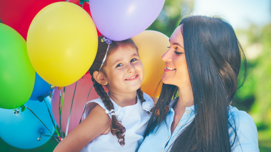 A mother smiles at the daughter in her amrs; the daughter smiles while holding a colorful assortment of balloons. 