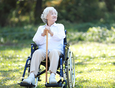 A woman in a wheel chair holds a cane while sitting in the sun, looking up hopefully.