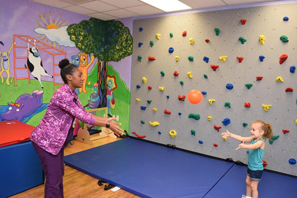 A child and therapist play with a ball in a brightly colored room with a rock wall.