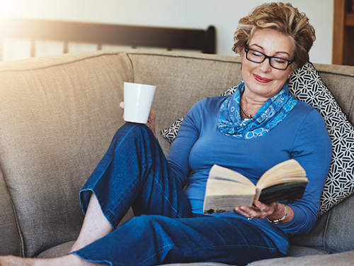 A woman leisurely reads a book on the couch, with coffee in hand.
